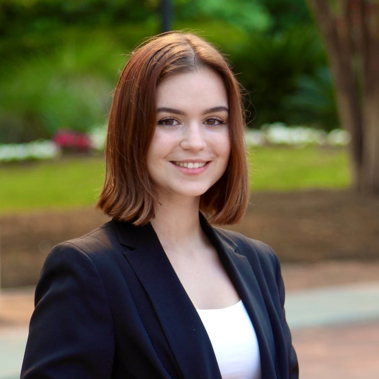 Alexa Scalchunes, Fall 2021 Outstanding Senior Scholar. Student is wearing a black blazer with a white camisole top underneath. Student has shoulder length auburn hair and is smiling. Background of photo is outside with pink and white blurred flowers.