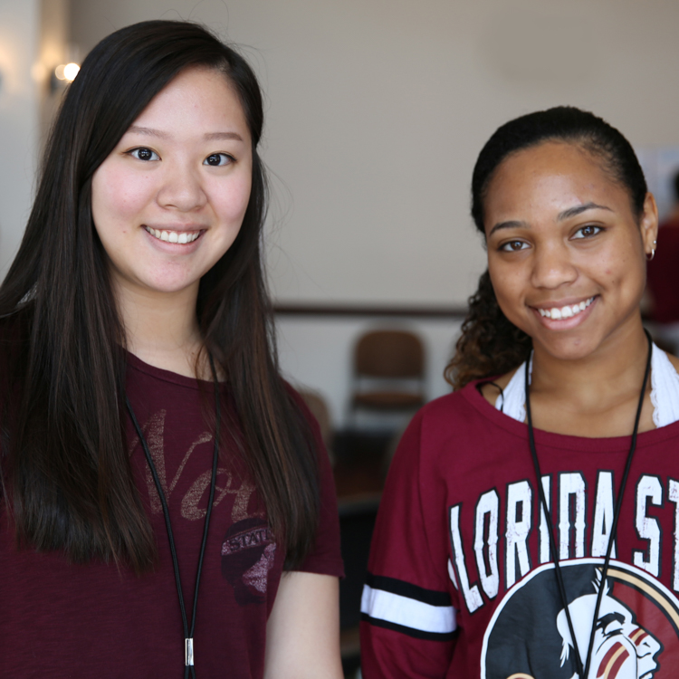 Two female honors students. Student on left is of Asian descent and has long dark brown hair and garnet shirt. She is smiling. Student on right is an African American female with hair pulled back into ponytail. She is wearing an FSU athletic looking shirt with the Seminole logo on the front. She is smiling. Background shows that students are in the Great Hall in the HSF Building on campus.