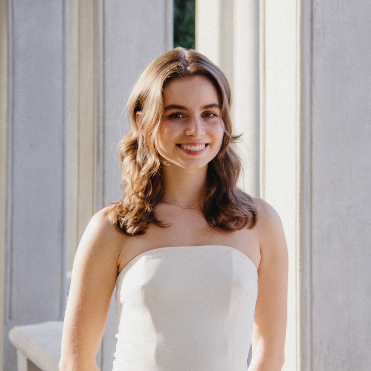 Aryanna Clark, Fall 2021 Outstanding Senior Scholar-Student has long wavy brown hair and is smiling. She is wearing a sleeveless white top with shoulders showing. The background includes different shades of white and gray. 