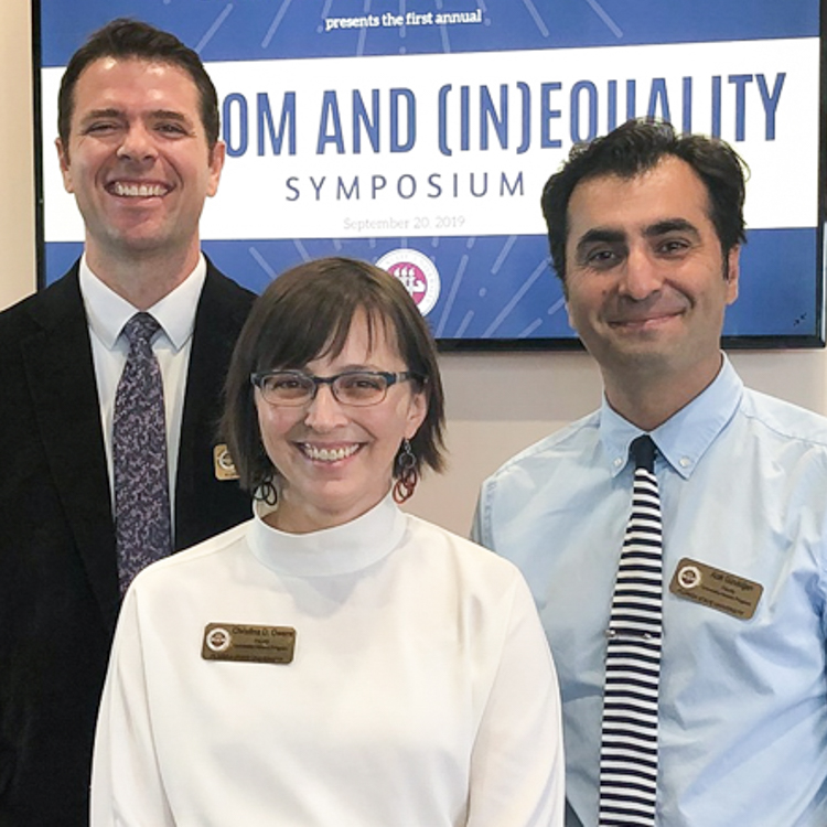 Three core honors faculty. Far left is tall man in suite with tie. In the center is female faculty in white mock turtleneck and glasses with name badge. Male faculty member on right is wearing a light blue buttoned down shirt with tie. He has dark short hair. All three are smiling for the camera. From left to right: Dr. Ross Moret, Dr. Christina Owens, and Dr. Azat Gundogan.