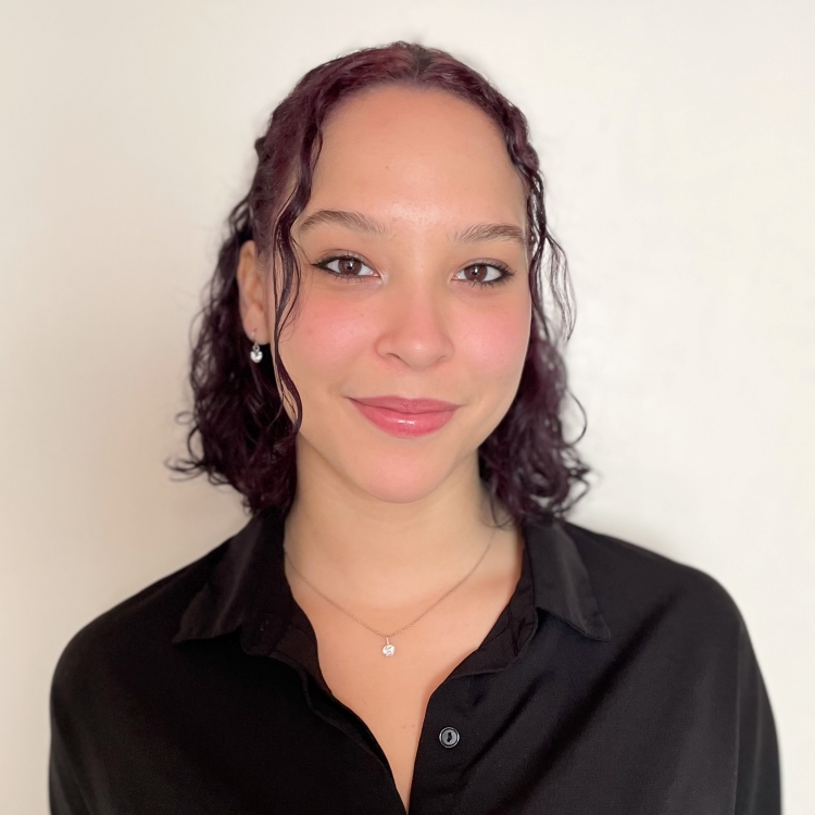 Photo of Mariella Soto, Fall 2021 Outstanding Senior Scholar. Mariella has shoulder length dark wavy hair, pulled back with a few fringe pieces on the sides of her face. She is wearing a black blouse, with her arms behind her back, sleeves folled up some. She has small diamond dangle earrings and a small diamond on a necklace. She is smiling.