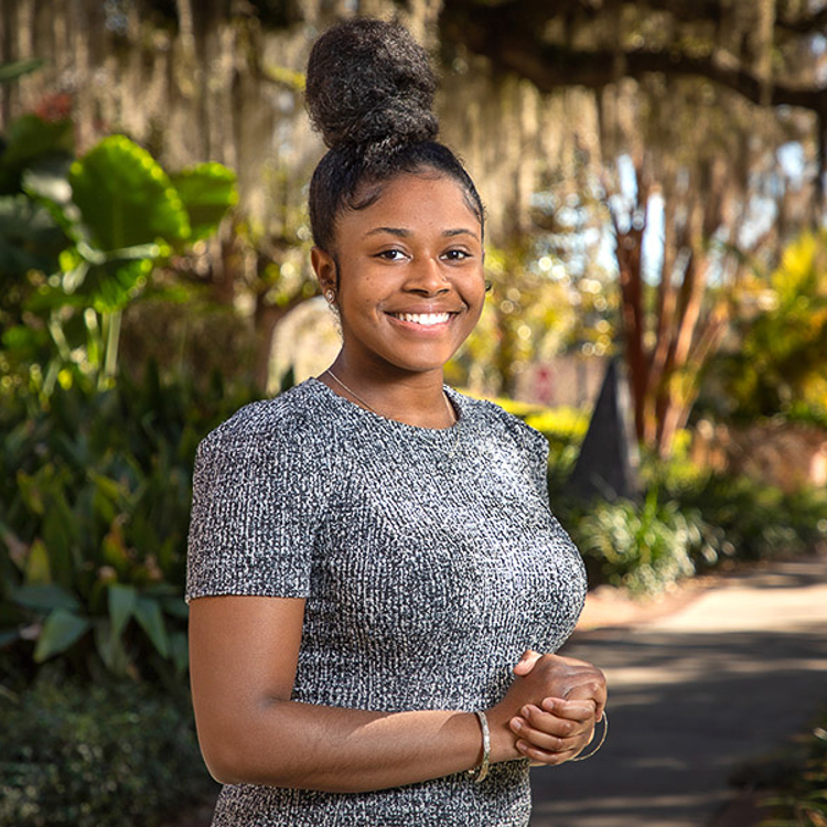 Ni'A Landon is wearing a salt and pepper short sleeved top and her hair is in a tall bun on top of her head. She is standing on a path outside the Westcott Building. Live oak tree with moss.