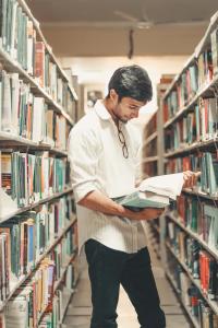 Young male student standing in library with bookshelves on either side of him. He has short dark hair and is wearing a white shirt. He is holding a couple books, one of which is opened and he is reading from it. Photo Credit: Photo by <a href="https://unsplash.com/@dollargill?utm_source=unsplash&utm_medium=referral&utm_content=creditCopyText">Dollar Gill</a> on <a href="https://unsplash.com/s/photos/college-students?utm_source=unsplash&utm_medium=referral&utm_content=creditCopyText">Unsplash</a>   