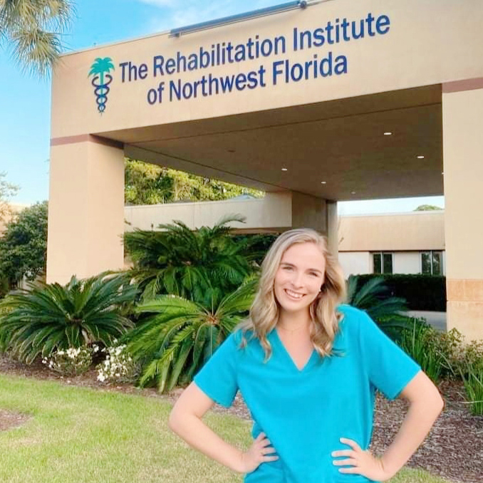 Honors in the Major Alumna, Ashley Archer-Ashley is wearing turquoise scrubs and is standing in front of her place of employment as a Speech Language Pathologist Clinical Fellow.