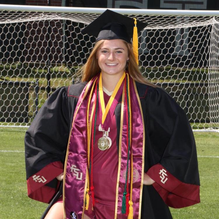 "Honors Alum, Kristina Lynch in full graduation regalia in front of soccer goal."