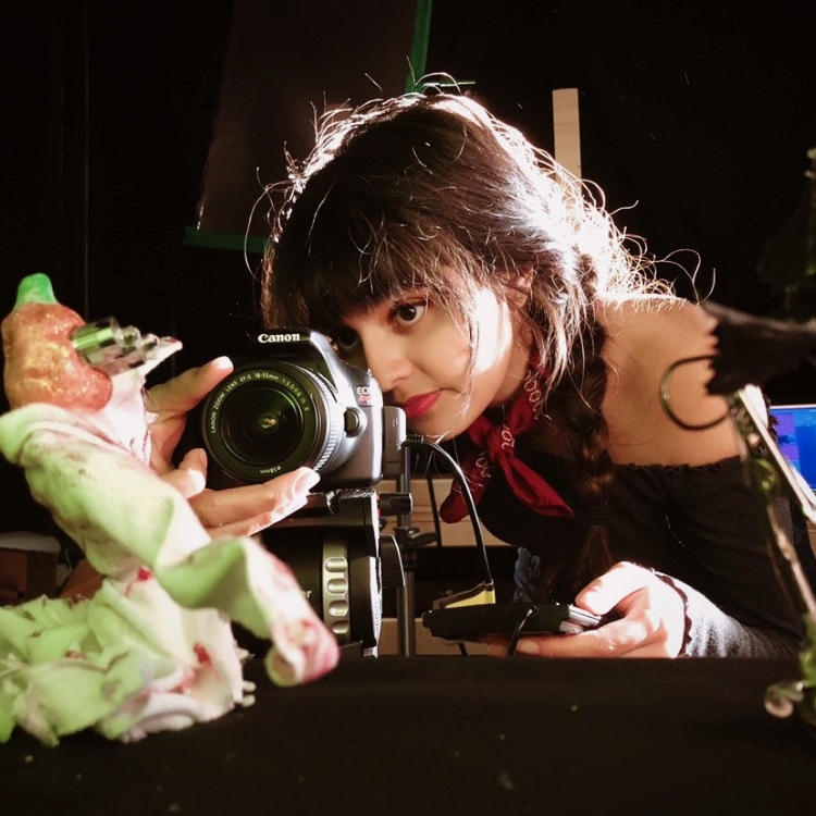 Student is looking through camera lens at subject of photograph. Background is dark and student's hair is illuminated by back lighting. Student has dark hair with bangs, with hair in braids. She is wearing off-the-shoulder black top with red bandana tied around her neck.Honors in the Major Student-Anisha Gupta-Film-Spring 2020