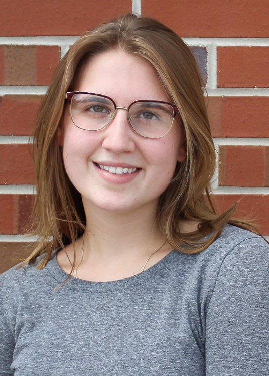 Student has shoulder length light brown hair. She is wearing a long sleeved heather gray t-shirt and glasses. She is standing in front of a brick wall and smiling.
