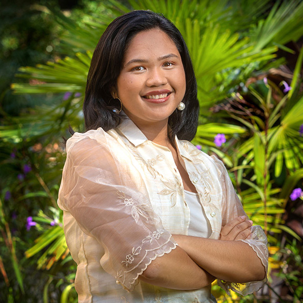 Christelle Bucag-student is wearing cream lacy dress and standing in front of palmetto plants. She is Filipino with dark hair and medium complexion. She is smiling.