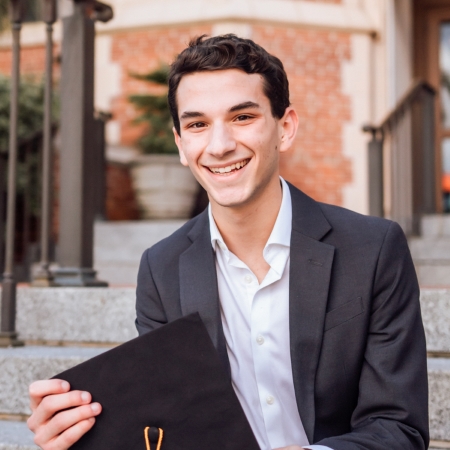 Image of Matthew Harris, Honors in the Major Student and Outstanding Senior Scholar, Spring 2021. Student has short, dark hair, is wearing a dark suit with buttoned down shirt. He is smiling. Sitting on steps to Westcott Building with his graduation mortar board in hand.