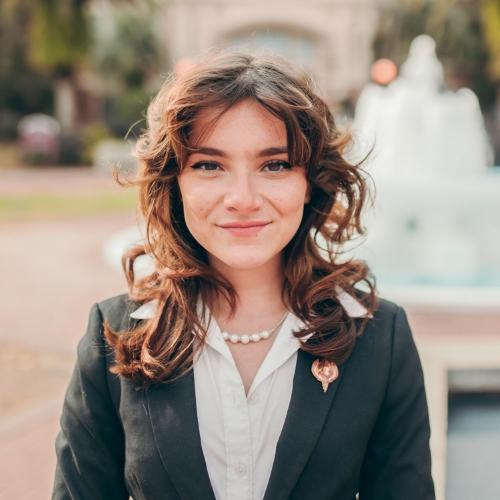 Image of Aliyah Hurt, Honors in the Major Student and Outstanding Senior Scholar Summer 2021. Student has long brown wavy hair. She is wearing a white buttoned down shirt with dark blazer. She is wearing an Outstanding Senior Scholar lapel pin and pearl necklace.