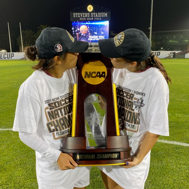 Two women's soccer players kiss the 2021 National Championship trophy. They are wearing white national champions t-shirts and dark ball caps. They are on the field after the game.