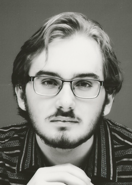 Black and White Photo of Student. He appears to have light brown hair with some blondish highlights. He has a dark short beard and mustache. He is wearing glasses. He has on a artistic striped shirt and his hands are cupped in the bottom center of the photo. 