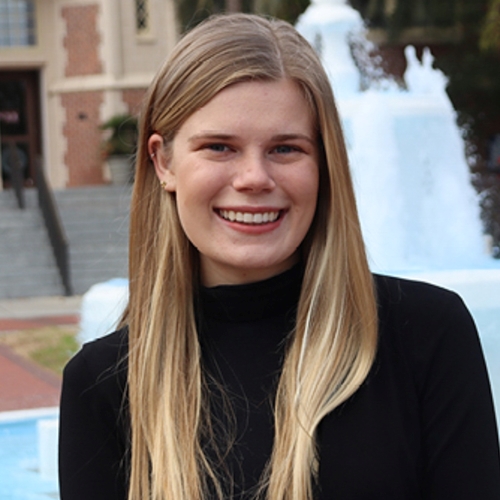 Maddy Johnson, Honors in the Major Student. She has long blonde hair and a black turtleneck shirt. She is sitting in front of the FSU Westcott Building fountain. She is smiling.