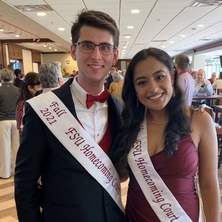 Student on left has short dark brown hair and glasses. He is wearing a black tuxedo and garnet bowtie. He is also wearing a white sash with the words Fall 2021 FSU Homecoming Court. Student on right is wearing a garnet formal full length gown with same white sash. Both are smiling. They are inside with lots of people in the background.