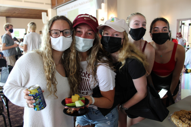 5 FSU University Honors Female Students with masks. Students are wearing casual clothes, including shorts. Student #1 (far left) has long blonde hair and glasses, and is holding a canned drink; Student #2 is wearing a garnet FSU cap, has long light brown hair and is holding a plate of cut melon fruit. Student #3 has long blonde hair and is wearing a white baseball cap with an embroidered pineapple and is wearing a black short-sleeved shirt. Student #4 has blonde hair in a tight ponytail and is wearing a garnet tanktop. Student #5 is has dark brown hair in a pony tail. She is wearing a red tank crop top and dark shorts. She is holding her phone. 