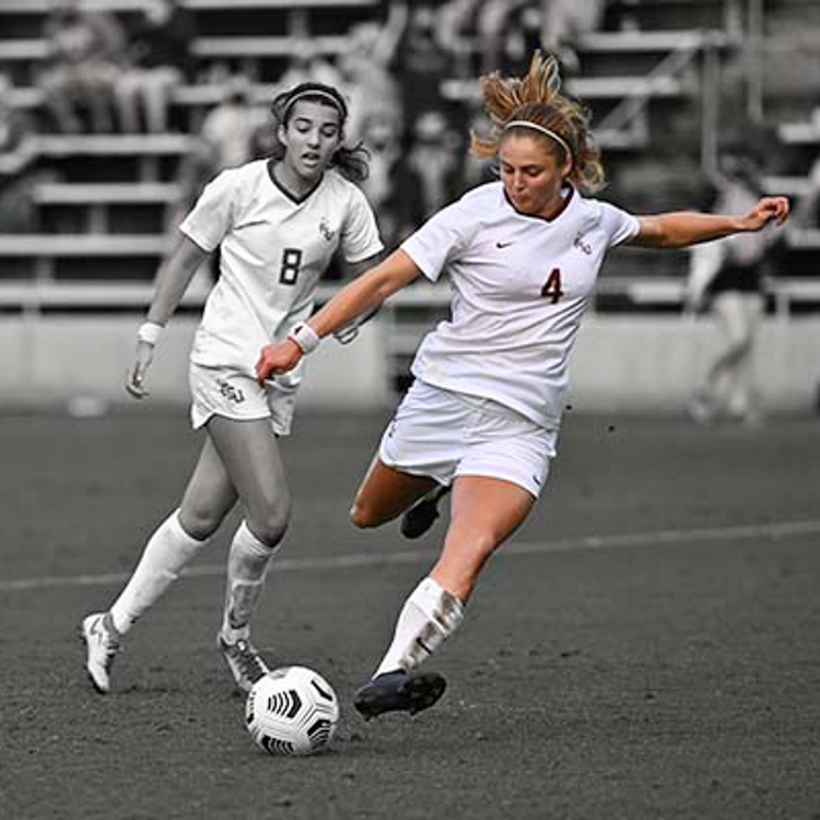 Two FSU Women's Soccer players. Kristina Lunch (on the right) is preparing to kick the ball. Photo is in black and white, but Kristina Lynch alone is in color. White soccer uniform. Photo credit: FSU Athletics. Link to College of Health & Human Sciences full write up on graduate student, Kristina Lynch.
