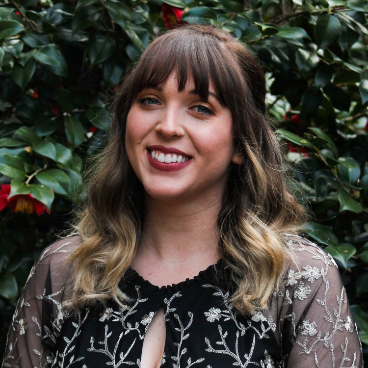 Photo of Megan Wright, Fall 2021 Outstanding Senior Scholar. Megan has brown hair with blonde highlights, also bangs. She is wearing a black dress with embroidered white flowers and sheer sleeves. She is standing in front of a camelia bush with red flowers. Student is smiling.