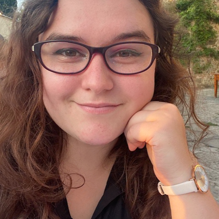 Image of Emily Pacenti, Honors in the Major Student, who completed an HITM thesis project. Part of the Student-Mentor Pair Profile Page. Student has long wavy brown hair and is wearing glasses. She is resting her chin on her left fist. She is smiling.
