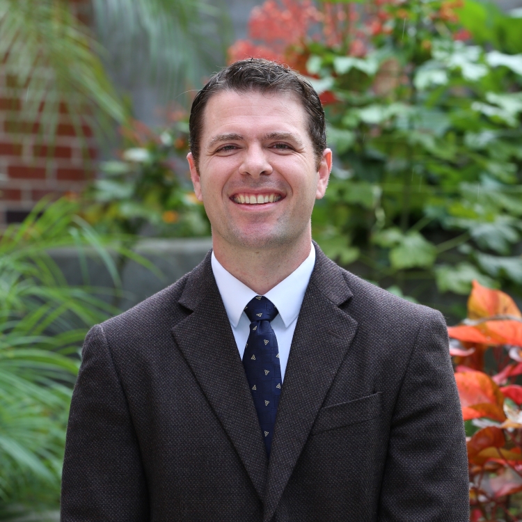 Dr. Ross Moret-Wearing dark suit jacket, dark tie and white shirt. Foliage in the background. He is smiling.