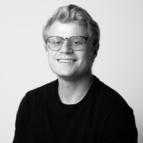 Image of Honors in the Major Student, Nicholas Tracanna. Black and white photo of student with blond hair, wearing dark shirt and glasses, smiling.